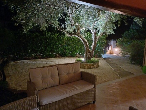 A wicker sofa with a cushion, a tree in a planter, and a paved area with a wall and greenery in the background.