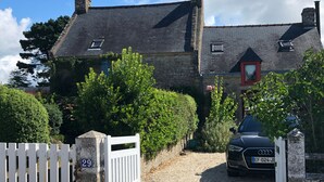 Een stenen huis met een grindpad, een wit hek en een geparkeerde auto.