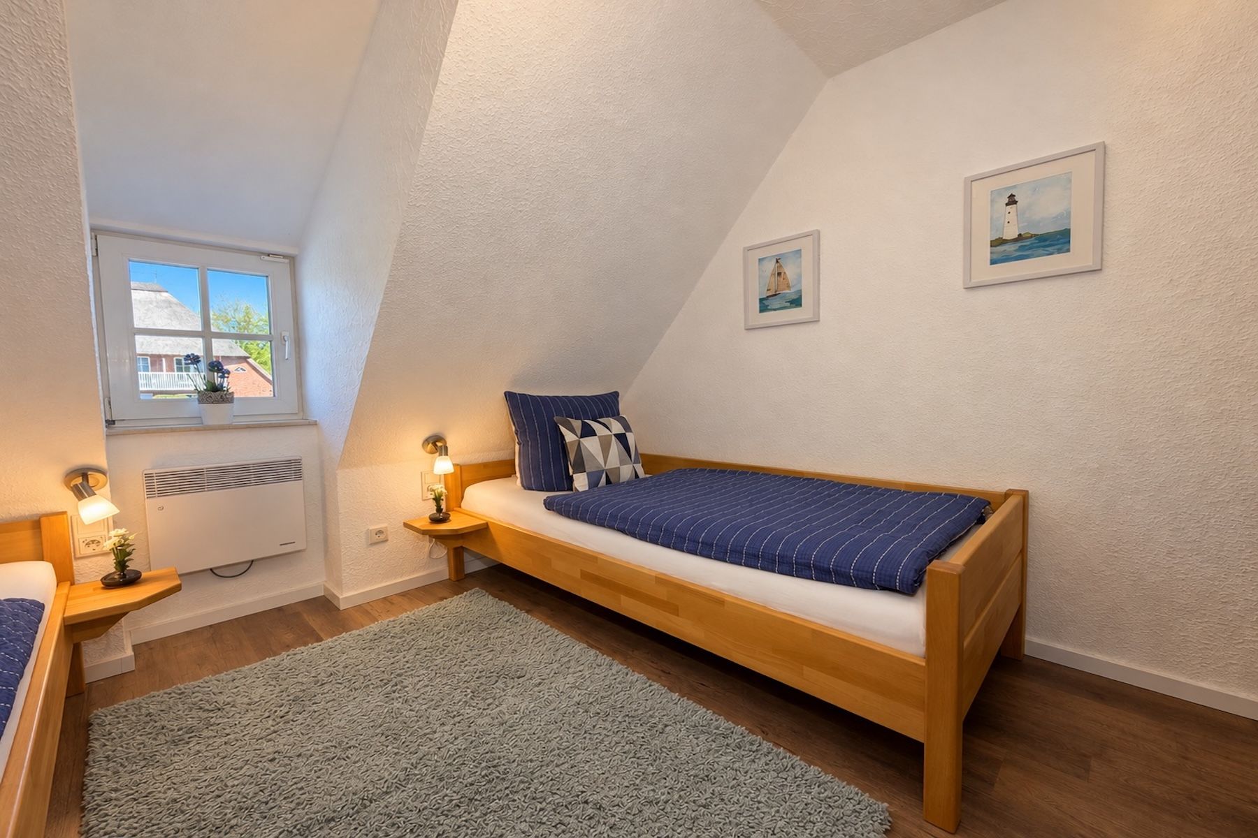Et soveværelse med skrå vægge, en seng med blå-stribet dynebetræk, et lille natbord med en lampe, et vindue med udsigt til et hus, og to billeder hængende på væggen.