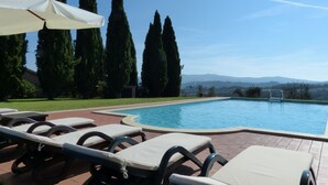Un área junto a la piscina con sillones de descanso y sombrillas, rodeada de altos árboles y vista a colinas distantes.
