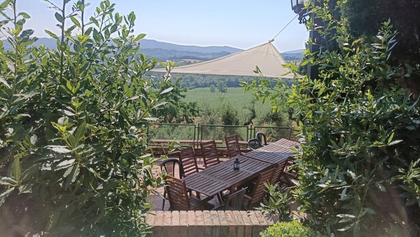 Una terraza con mesa y sillas, rodeada de vegetación, con vistas a un paisaje con colinas.