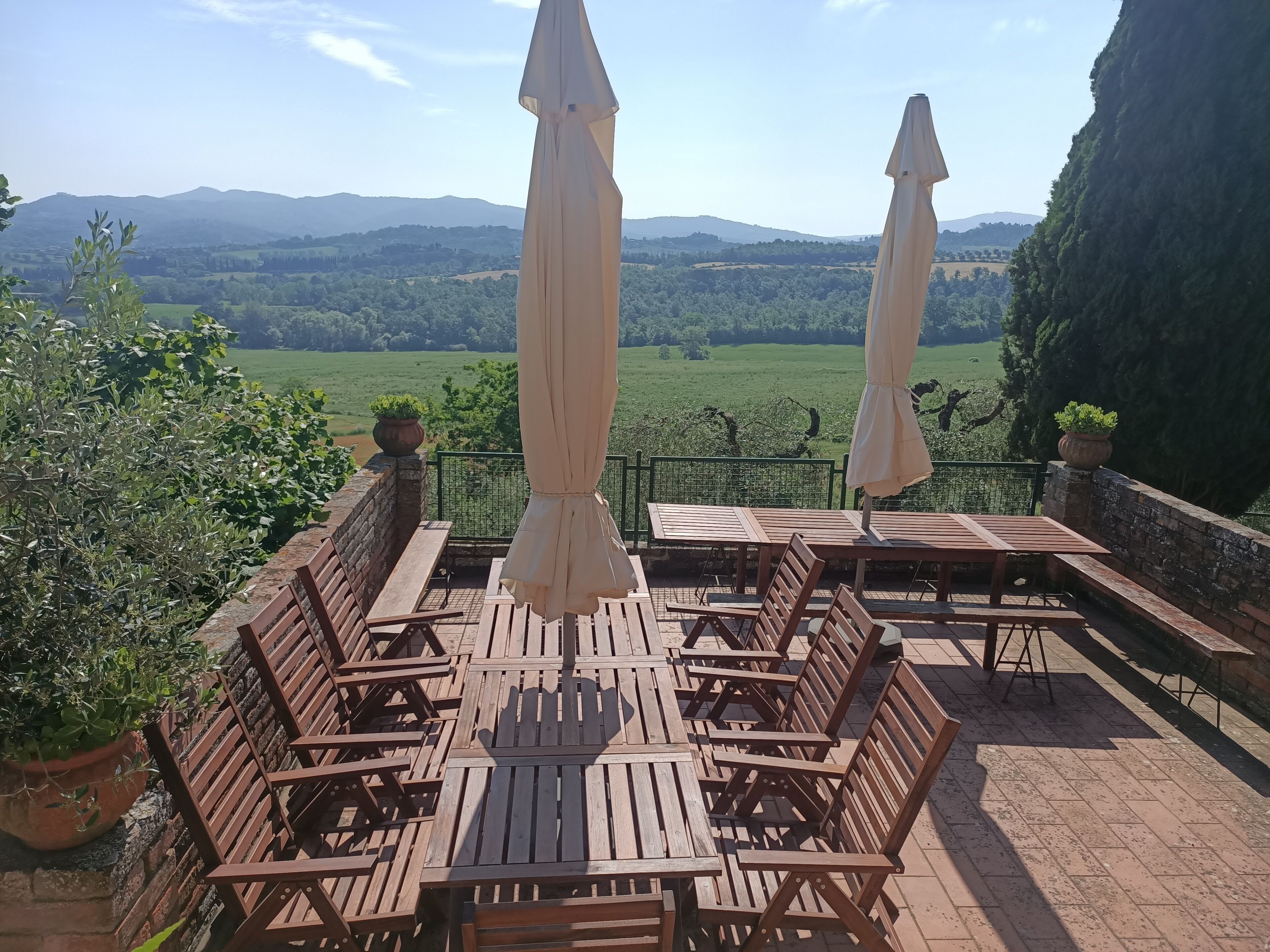 An outdoor seating area with wooden tables and chairs, two beige umbrellas, and a view of green fields and mountains in the background.