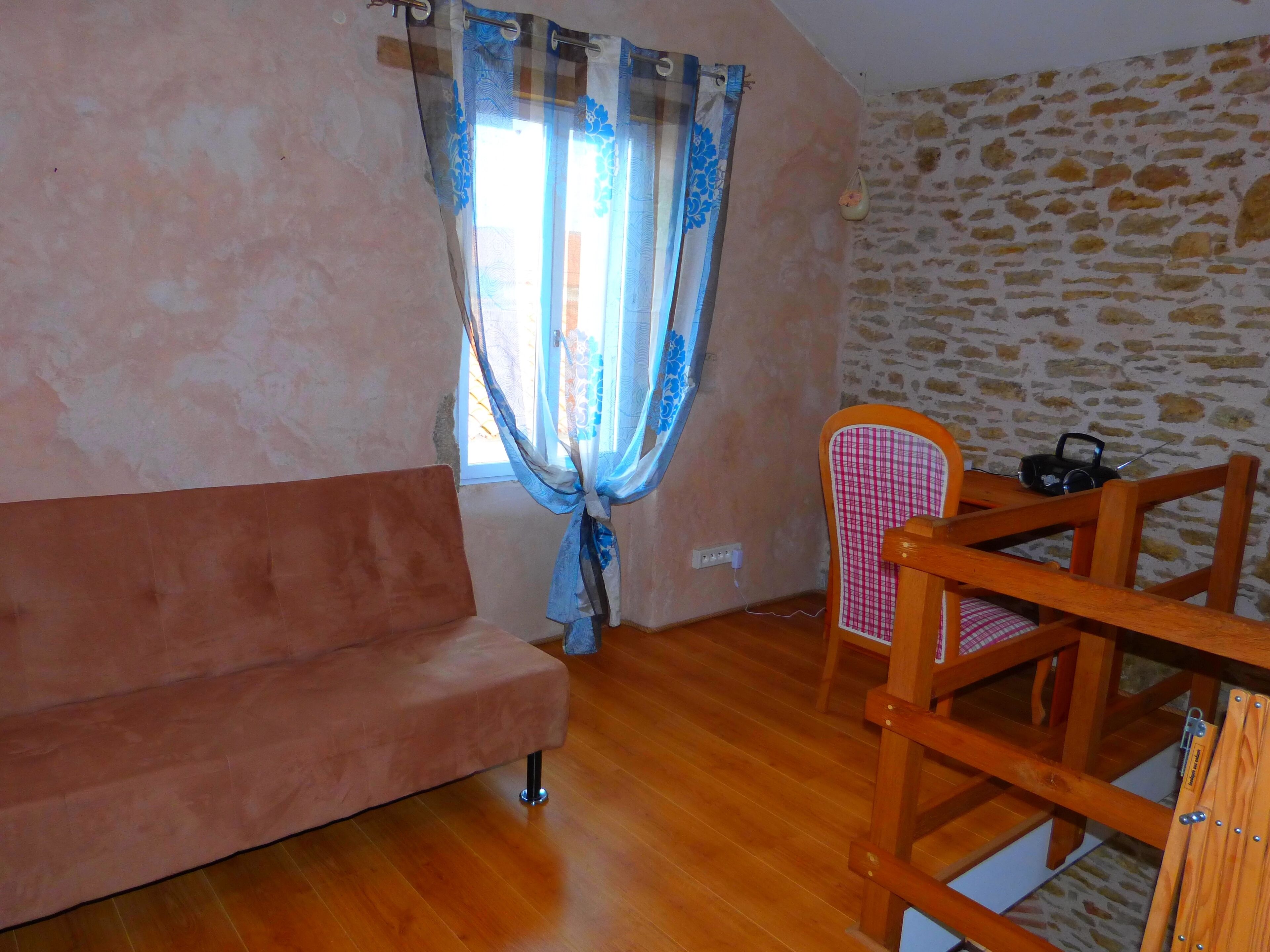 Une pièce avec un plancher en bois, un canapé rose, une chaise avec un coussin à carreaux, une table avec une radio et une fenêtre ornée de rideaux à motifs.