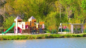 A playground with slides and climbing structures near a body of water, surrounded by trees.