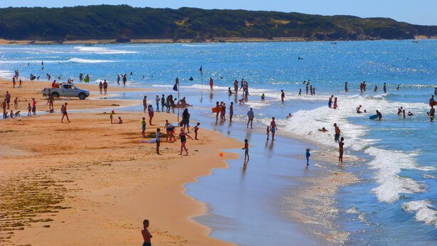A crowded beach with people engaging in various activities, a lifeguard station, and a sailboat in the distance.