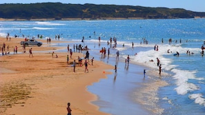 Una playa concurrida donde la gente participa en diversas actividades, una estación de salvavidas y un velero en la distancia.