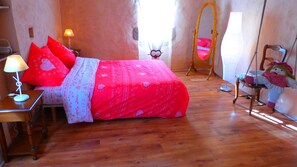Une chambre avec un plancher en bois, un lit recouvert d’une couvre-lit à motifs rouges et blancs, une table de chevet avec une lampe et un fauteuil suspendu.