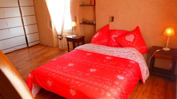 Un dormitorio con piso de madera, una cama con ropa de cama roja, una mesita de noche con una lámpara y un aparador con un espejo.