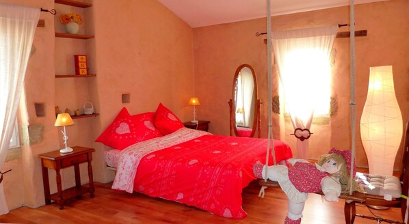 Une chambre à coucher comprenant un lit, une table de chevet, une lampe, un miroir, un fauteuil suspendu et une étagère avec des objets décoratifs.