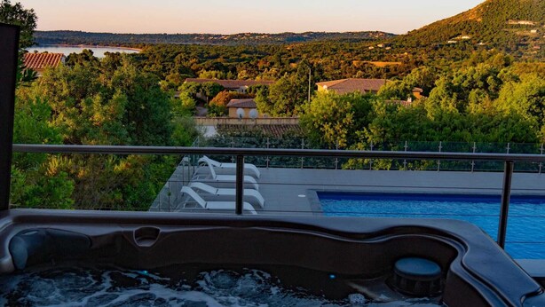 Un bain à remous avec une piscine, entouré d’une végétation luxuriante et offrant une vue sur une colline parsemée de bâtiments.