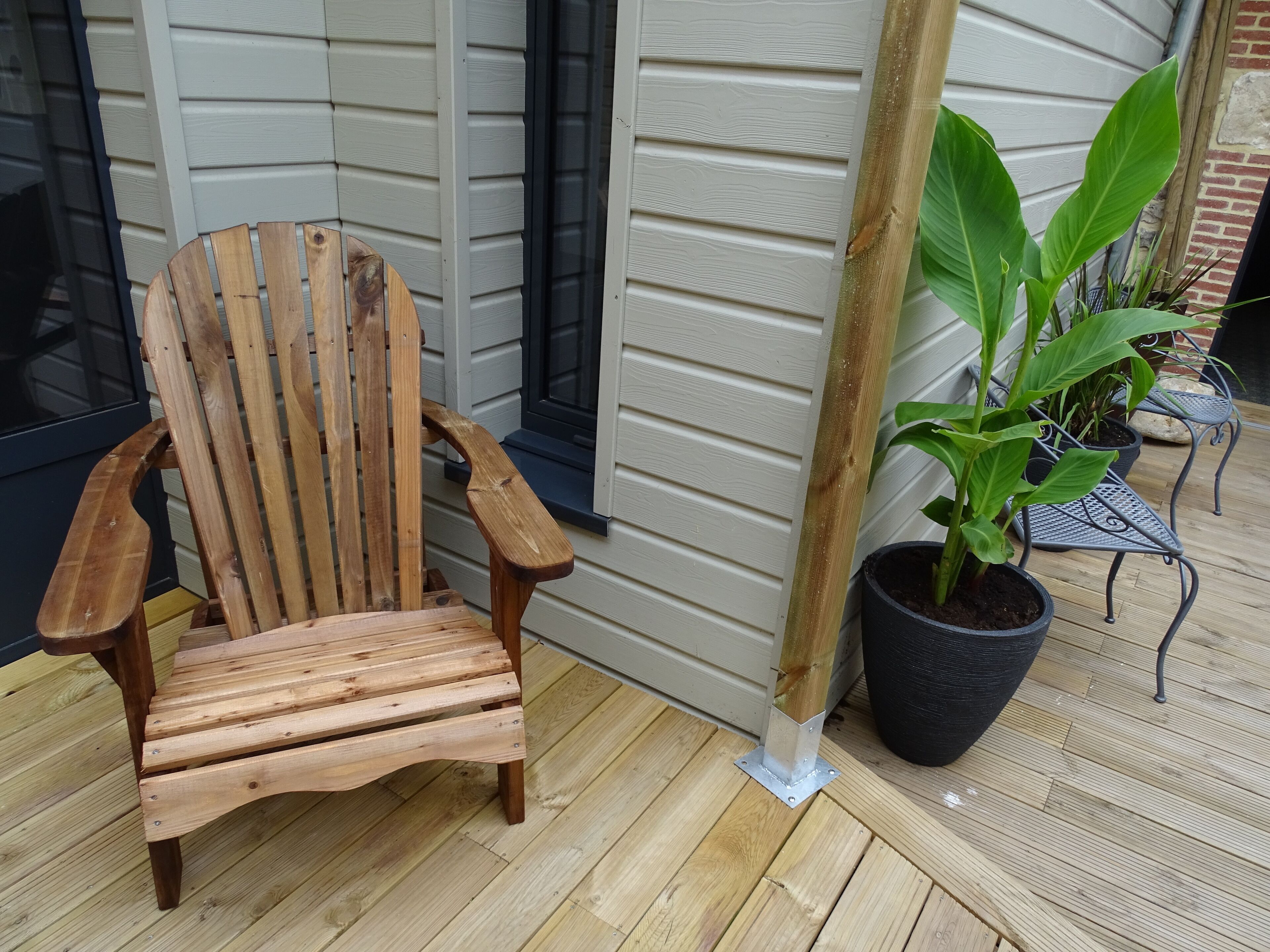 En trästol i Adirondack-stil, en krukväxt och en bänk i metall på ett trädäck.