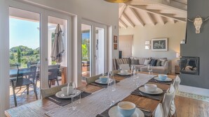 Un coin repas avec une longue table dressée pour six personnes, un canapé en arrière-plan et une vue sur la verdure à l’extérieur.