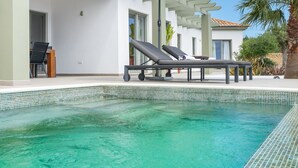 Une piscine avec un bord carrelé, une chaise longue et un parasol.