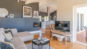 Un salon moderne avec un canapé gris, une table basse noire et un téléviseur. On aperçoit une cuisine au fond.