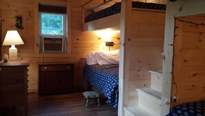 Een houten kamer met een stapelbed, een bureau met lamp, een klein krukje en een raam met uitzicht op bomen.