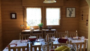 Spiseområde i en træhytte med et bord dækket til fire personer, to vinduer, en hængende lampe og en vægmonteret dekoration af et hjortehoved.