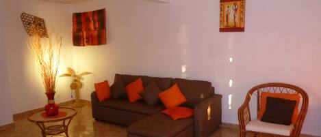 A living room with a brown sectional sofa, a small round table with a vase, a wicker chair, and artwork on the walls.