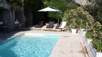 Une piscine entourée de verdure et de murs de pierre, avec des chaises longues et des parasols.