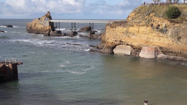 Une plage avec un rivage rocheux, une personne assise sur le sable et un pont en arrière-plan.