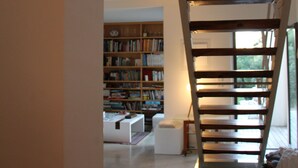 Eine Treppe mit Holzbodenstufen führt zu einer Zwischenebene, wo sich ein Bücherregal voller Bücher und ein weißer Hocker befinden.