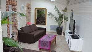 Un salón con un sofá marrón, una mesita con mantel rosa, una planta en maceta, un televisor sobre un mueble bajo y una pared de ladrillo con una ventana.