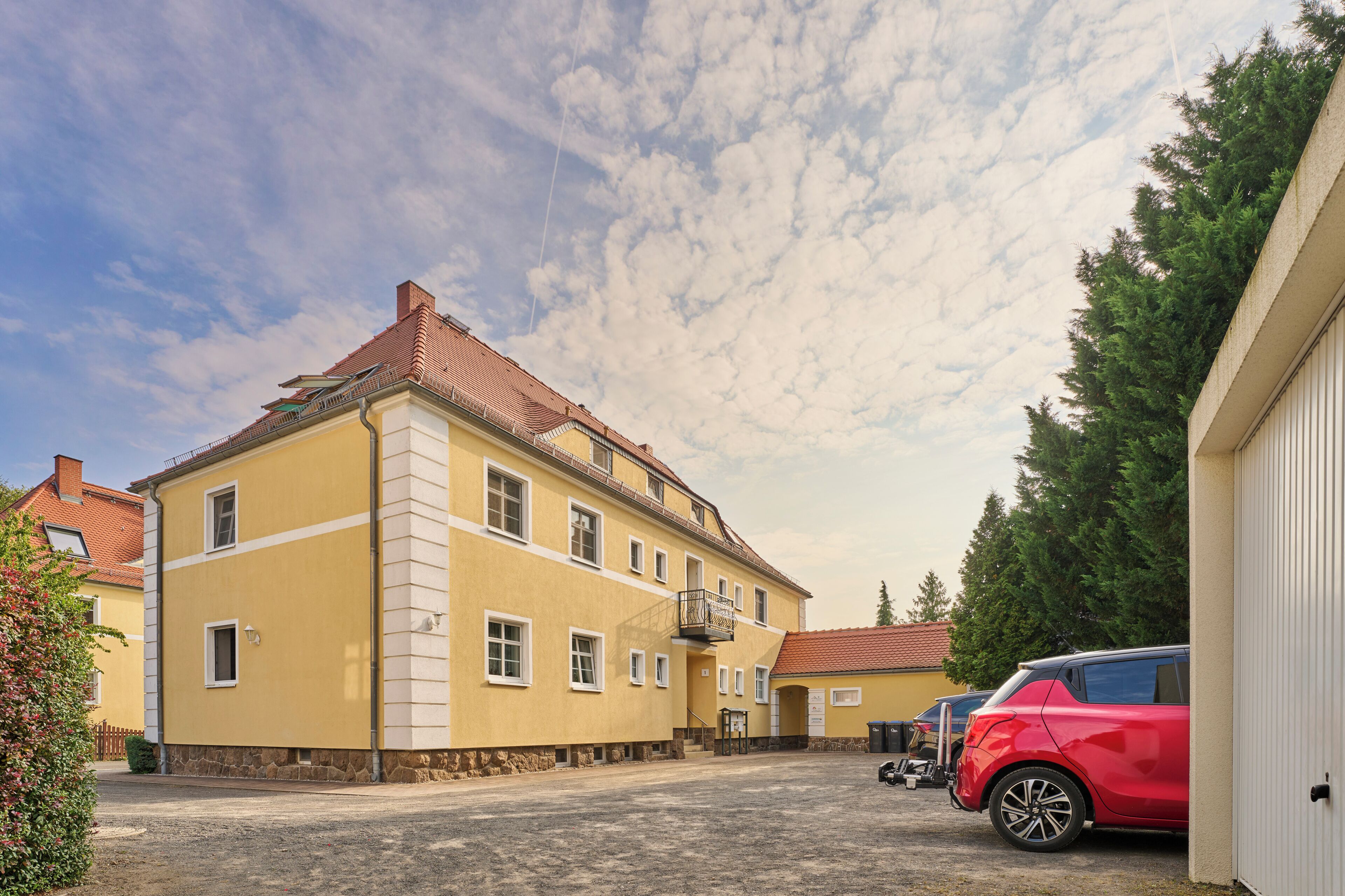 Un bâtiment jaune avec un toit rouge, un escalier et une voiture rouge garée devant.