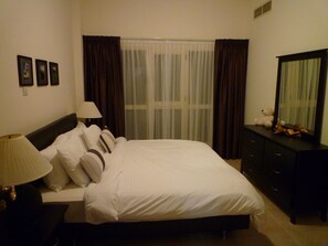 A neatly made bed with white linens and a dark headboard. There is a lamp on a bedside table to the left. A dresser with a mirror is to the right of the bed. - Townhouse with pool, near the beach, right at the golf course (Ras al Khaimah)