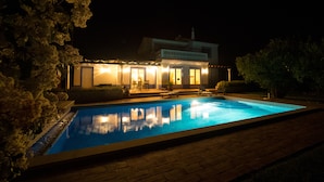 Vista nocturna de una casa con piscina, un patio con mobiliario exterior y una entrada bien iluminada.
