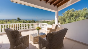 Un balcón con mobiliario de mimbre, una mesa con vasos y vistas al mar y la vegetación.