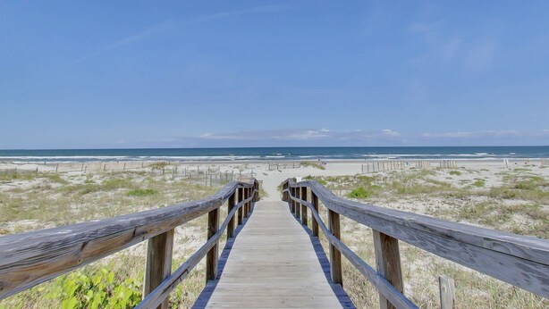 Ein hölzerner Steg führt zu einem Strand mit Dünen und wolkenlosem, blauem Himmel.