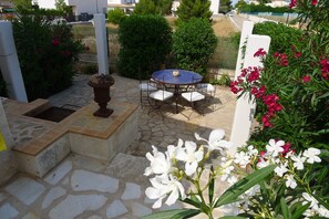 Un patio de piedra con una mesa redonda y sillas, rodeado de vegetación y flores.