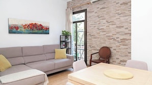 Ein Wohnzimmer mit einer Couch, einem Esstisch und einem Stuhl. An der Wand hängt ein Gemälde und das Fenster bietet Blick auf grüne Vegetation.