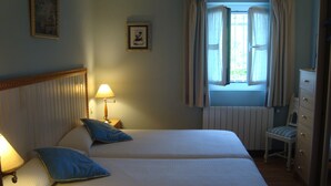 Una cama de matrimonio con dosel, dos almohadas, una lámpara de noche, una ventana con cortinas, un radiador y una silla con cojín a rayas.