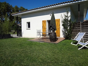 A white building with a tiled roof, a wooden door, and a small window. There is a table with chairs and a barrel in the yard.