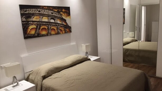 Un dormitori amb un llit, taules de nit, una làmpada, un mirall i un quadre del Colosseu.