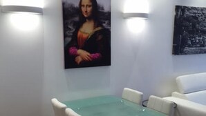 Zona de menjador amb taula de vidre i cadires blanques, un sofà blanc i il·luminació penjada a la paret.