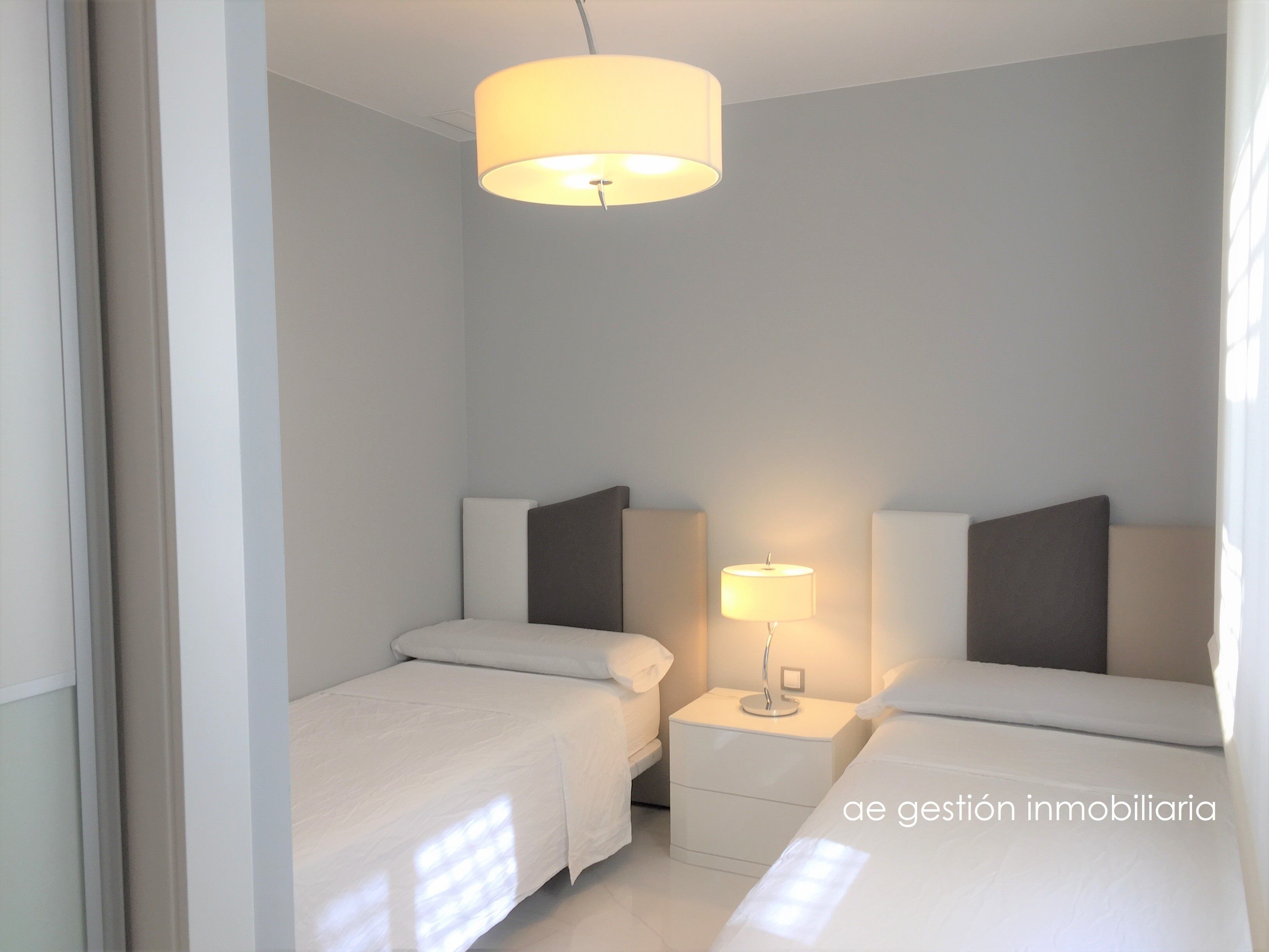 A hotel room with two beds, a nightstand with a lamp, and a ceiling light.