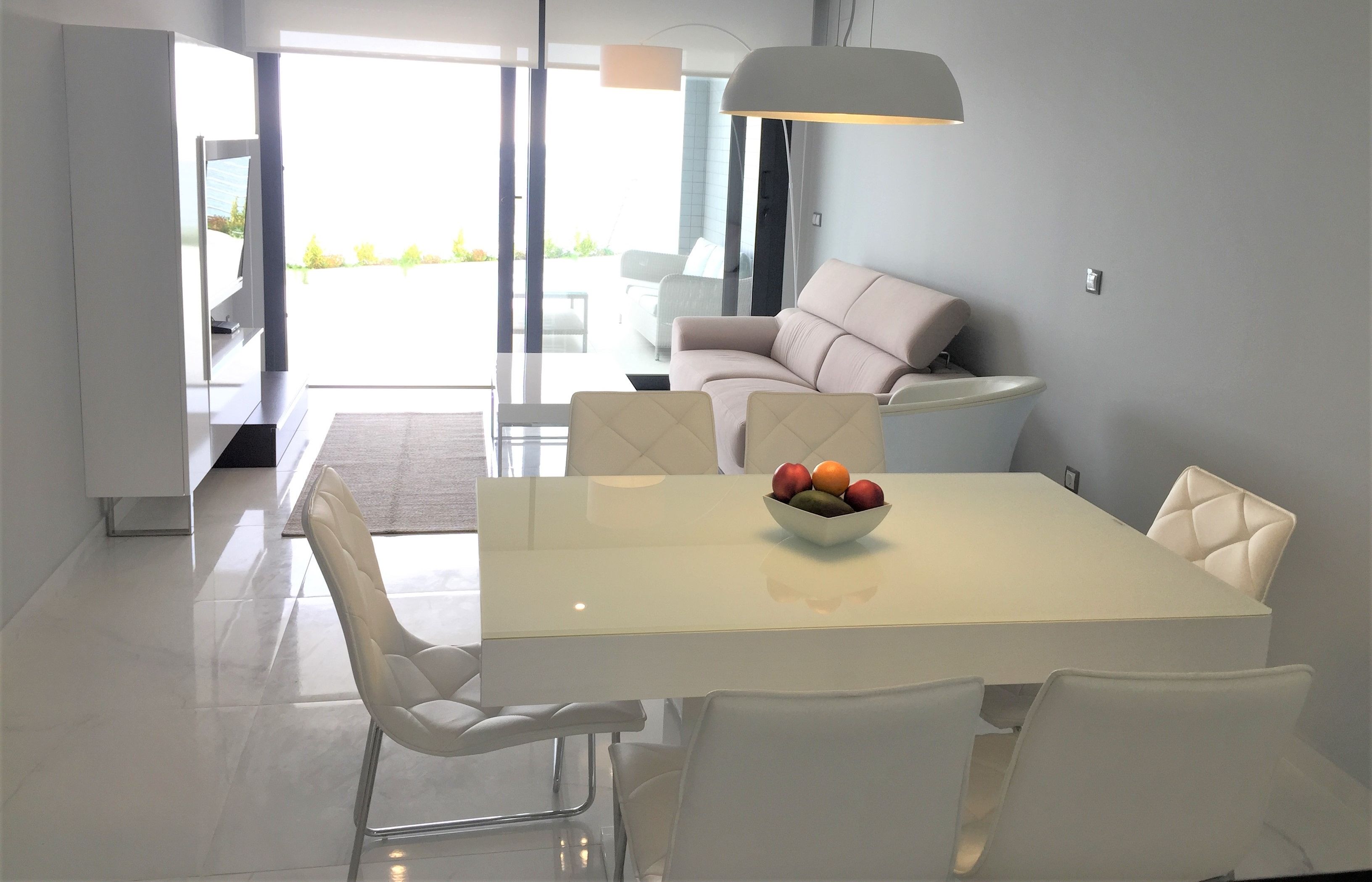 A modern dining area with a white table and chairs, a bowl of fruit, and a sofa in the background.