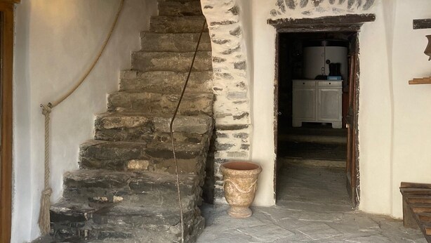 Uma escadaria de pedra leva a uma porta, com um grande vaso de terracota e um armário de madeira em um ambiente rústico.