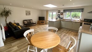 A modern kitchen with white cabinets, a dining area with a wooden table and white chairs, and a living room with a black sofa and a television. - Mill Lane Cottage, a pretty cottage in beautiful countryside, 4 miles from Bath. (Bath)