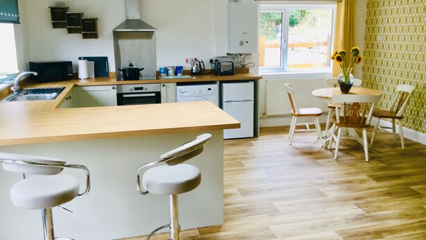 A modern kitchen with a central island, bar stools, a dining table with chairs, and a window with curtains. - Mill Lane Cottage, a pretty cottage in beautiful countryside, 4 miles from Bath. (Bath)