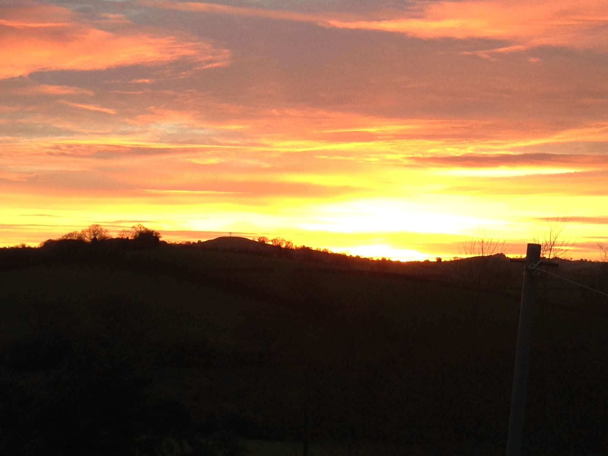 A sunset over a hilly landscape with a silhouette of trees and a utility pole.