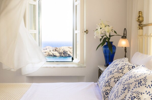 A bedroom with a bed, a vase with flowers, a lamp, and a view of the sea through an open window.