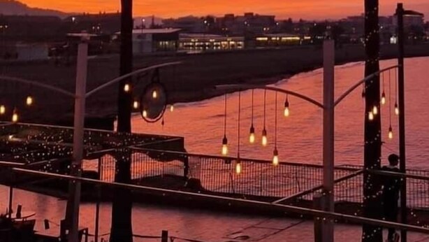 En restaurant ved vandet med udendørs siddepladser, palmer og udsigt til solnedgang.