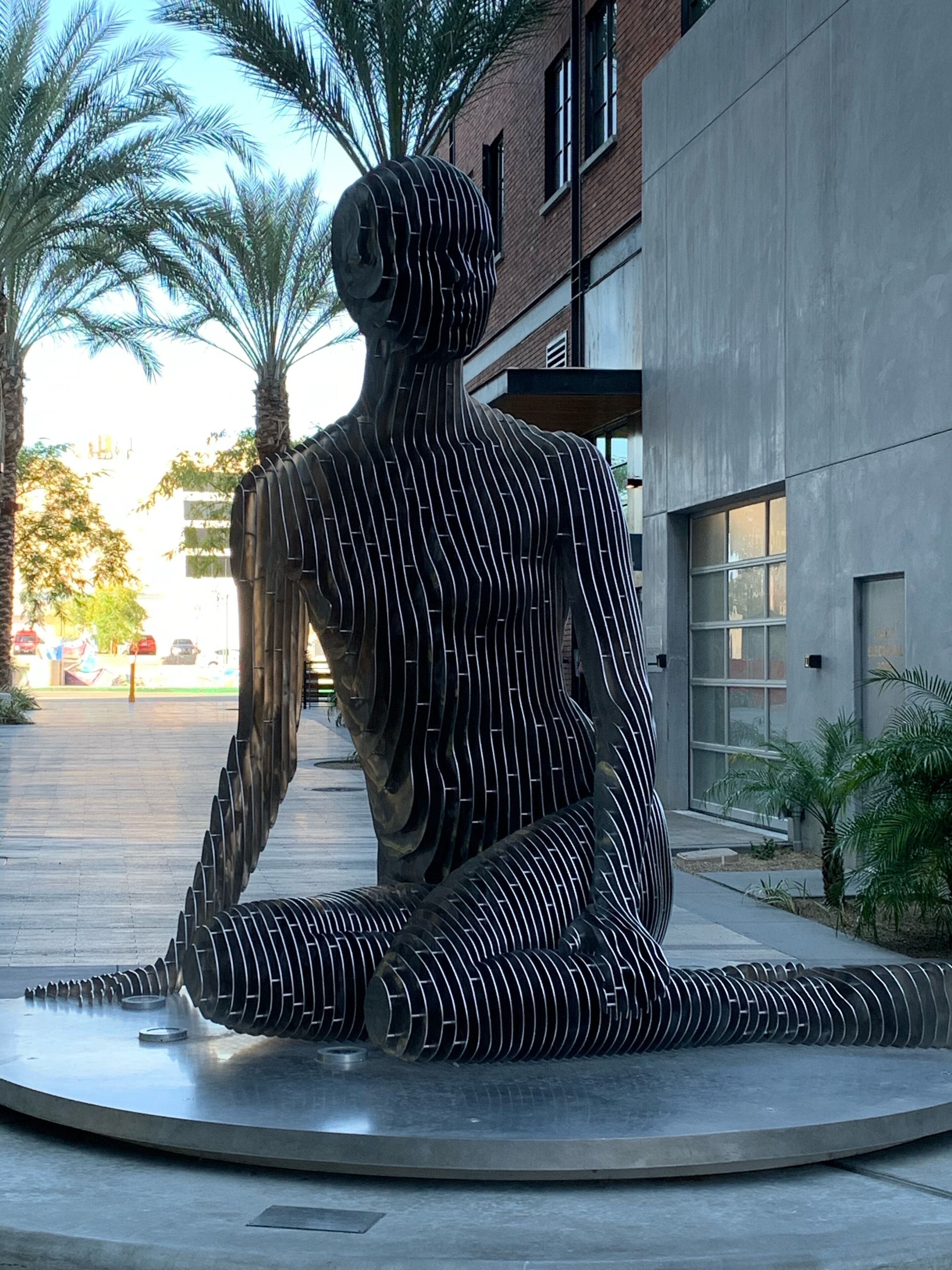 Une sculpture moderne représentant une figure assise, composée de lignes verticales, et située dans un environnement urbain, avec un bâtiment et des palmiers en arrière-plan.
