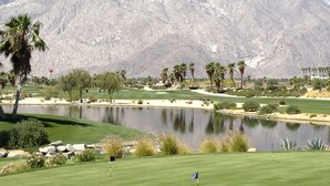 A golf course with a pond, palm trees, and mountains in the background. - Bright and Cheerful South Desert Home (Palm Desert)
