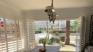 A dining area with a wooden table and benches, a chandelier, and a view of a desert landscape through open windows. - Bright and Cheerful South Desert Home (Palm Desert)