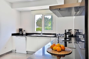 A modern kitchen with white cabinetry, a black countertop, a built-in dishwasher, a stainless steel range hood, and a window with a view of greenery.