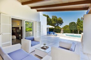A covered outdoor area with wicker furniture, a pool, and a view of trees and a clear sky.