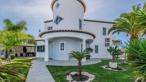 Une maison moderne à deux étages, dotée d’une façade incurvée, d’un patio couvert et d’un jardin bien entretenu avec des palmiers et un sentier.